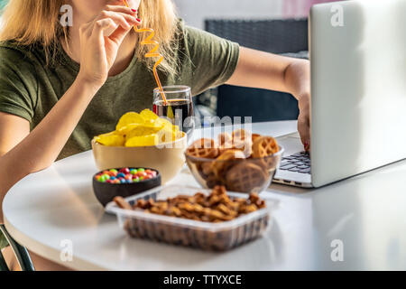 Ragazza lavora a un computer e si mangia il fast food. Cibo malsano: chips, cracker, candy, waffle, cola. Il cibo spazzatura, concetto. Foto Stock