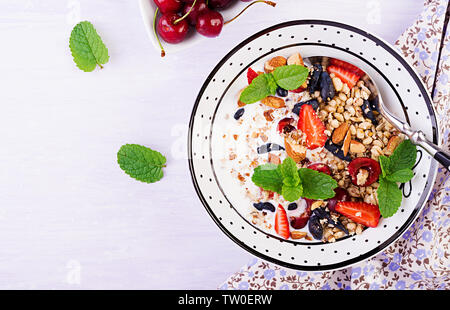 Una sana prima colazione - granola, fragole, ciliegia, caprifoglio berry, dadi e lo yogurt in una ciotola. Concetto vegetariano cibo. Vista superiore Foto Stock