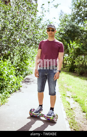 Giovane uomo moderno in denim shorts e borgogna t-shirt passeggiate in giro per la città su un hoverboard Foto Stock