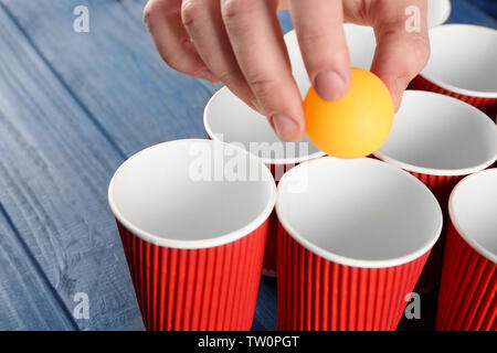 Giallo palla in mano maschio e bicchieri di carta disposti per giocare Pong della birra Foto Stock