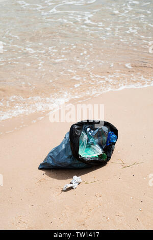 Vista dettagliata del grigio di un sacchetto di plastica con rifiuti raccolti sulla sabbia di una spiaggia solitaria, accanto all'acqua, in background Foto Stock