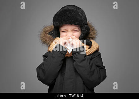 Carino piccolo ragazzo in vestiti caldi utilizzando un tessuto per soffiare il naso su sfondo grigio Foto Stock