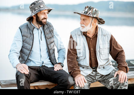 Nonno con Figlio seduto e parlare insieme sul banco di lavoro mentre la pesca sul lago di prima mattina Foto Stock