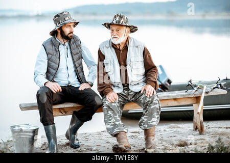 Nonno con Figlio seduto e parlare insieme sul banco di lavoro mentre la pesca sul lago di prima mattina Foto Stock