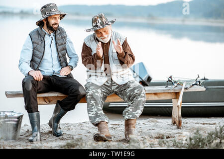 Nonno con Figlio seduto e parlare insieme sul banco di lavoro mentre la pesca sul lago di prima mattina Foto Stock