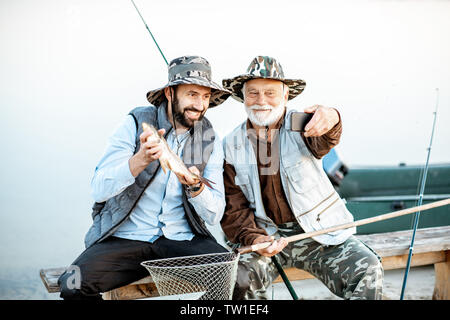 Nonno con figlio avendo divertimento, rendendo selfie foto insieme con il pesce appena pescato sul lago di prima mattina Foto Stock