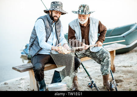 Felice nonno con figlio tenendo il pesce appena pescato seduti insieme vicino al lago di prima mattina Foto Stock