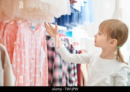 Vista laterale di funny girl in bianco maglione guardando il bellissimo abito rosa in negozio. Bella femmina bambino cercando per scegliere e acquistare nuovi vestiti in boutique. Concetto di vendita e shopping. Foto Stock