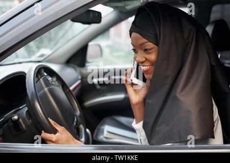 Elegante musulmani africani donna che indossa in nero hijab seduto in automobile, parla al telefono cellulare. Bella e giovane ragazza tenendo una mano sul volante, sorridente. Foto Stock