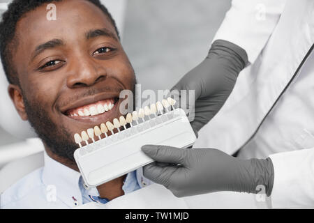 Giovane africano guardando la fotocamera e sorridere mentre dentista femmina mantenendo la gamma dei riempimenti in clinica. Felice paziente bianco godendo di uno splendido sorriso mentre la visita studio dentistico. Concetto di cura. Foto Stock