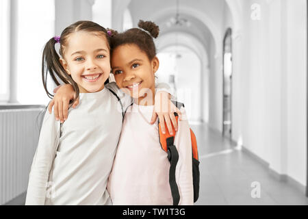 Ritratto di due migliori amici piccola scuola ragazze in piedi in corridoio, abbracciando ogni altro e sorridente. African e Caucasico ragazze che studiano insieme a scuola. Concetto di amicizia. Foto Stock