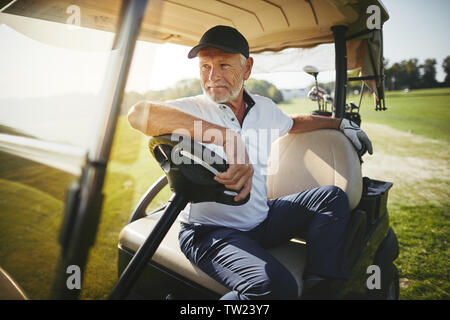 Senior sorridente uomo seduto in un golf cart a guardare il fairway durante la riproduzione di un round di golf in una giornata di sole Foto Stock