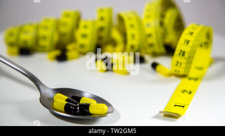 Cucchiaio con nero e giallo pillole e nastro di misurazione su sfondo bianco per rappresentare la dieta pillola industria. Mangiare sano e di integratori alimentari Foto Stock