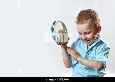 Allegro ragazzo in una maglietta blu tenendo un tamburello e sorridente su uno sfondo luminoso Foto Stock