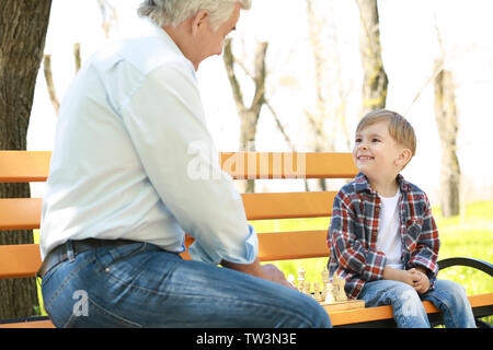 Carino piccolo ragazzo con il nonno gioca a scacchi sulla panchina nel parco di primavera Foto Stock