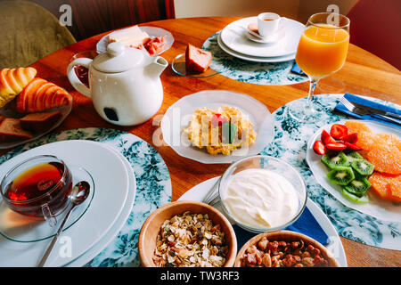 Tavolo per la colazione con una varietà di alimenti compresi i cereali, yogurt, uova strapazzate, frutta, croissant e bevande come tè, caffè e succo d'arancia. Foto Stock