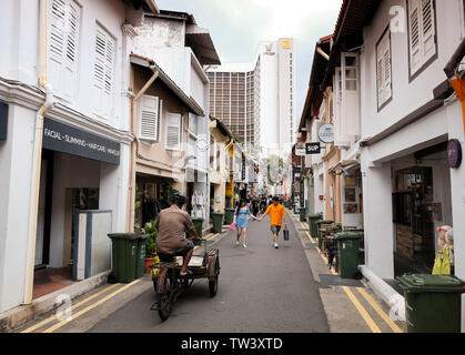 Un vecchio uomo cavalca un triciclo con una consegna di pacchi carrello collegato alla parte anteriore lungo Haji lane in Singapore. Foto Stock