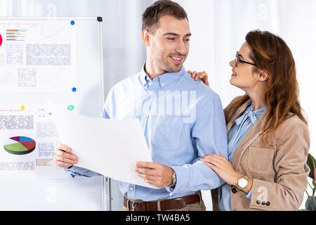 Donna allegra in bicchieri guardando uomo bello con carta bianca nei pressi di lavagna bianca con tabelle e grafici Foto Stock