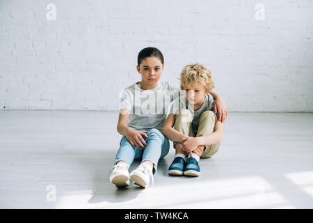 Triste sorella seduta sul pavimento e abbracciando il fratello Foto Stock