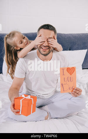 Uomo sorridente azienda confezione regalo e padri giorno greeting card mentre allegro figlia che copre la sua gli occhi con le mani Foto Stock