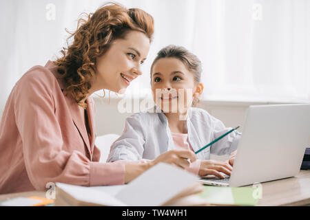 Felice madre rivolto verso lo schermo del notebook mentre aiuta la figlia a fare i compiti di scuola Foto Stock