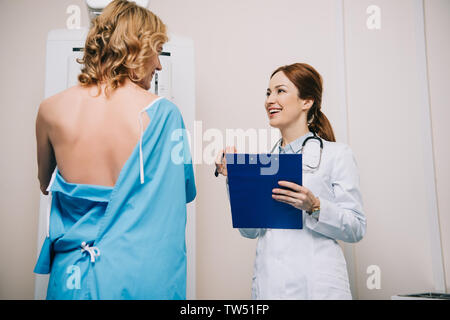 Sorridente radiologo azienda appunti mentre sta in piedi vicino al paziente facendo x-ray prova per mammografia Foto Stock