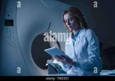 Medico grave guardando a tavoletta digitale con raggi x la diagnosi mentre è seduto vicino a CT scanner Foto Stock