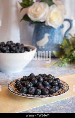 Mirtilli freschi raccolti su un piatto e in una ciotola bianca con tovaglie e fiori sullo sfondo presentati in ambiente luminoso e luminoso cucina. Foto Stock