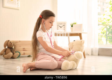 Adorabile bambina esaminando orsacchiotto con stetoscopio rosa a casa Foto Stock