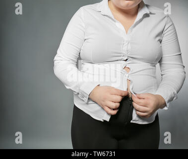 Il sovrappeso donna cercando di pulsante in alto la sua camicia su sfondo grigio. La perdita di peso concept Foto Stock