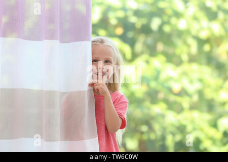 Incantevole piccola bambina che si nasconde dietro la tenda a casa Foto Stock