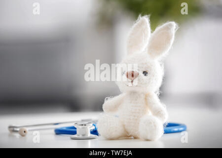 Simpatico coniglietto di maglia e uno stetoscopio sul tavolo contro sfondo sfocato. Concetto di visitare i bambini del medico Foto Stock