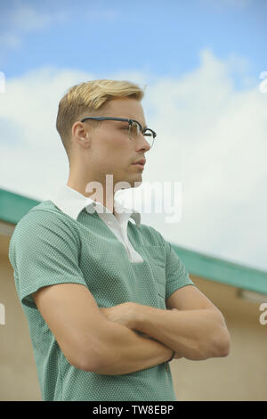 Un maschio bianco caucasico che indossa una polo verde anni '60 e occhiali vintage, braccia piegate guardando via. Dietro di lui c'è un cielo blu. Foto Stock