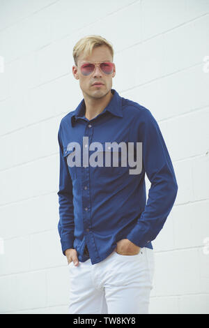 Un modello maschio caucasico biondo che si posa in una camicia d'epoca blu 70s, posando all'aperto su uno sfondo bianco parete. Foto Stock