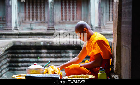 Angkor Wat, UNESCO, Siem Reap, Cambogia - Agosto 2016: un arancione-derubato monaco seduto al corridoio's edge e preparare il cibo con un tempio sfocata b Foto Stock