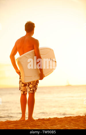 Surfer uomo sulla spiaggia al tramonto di surf holding bodyboard. Montare il corpo maschio surfer guy godersi il tramonto e bodyboarding sulle vacanze estive vacanze sulla spiaggia tropicale. Kaanapali Beach, Maui, Hawaii. Foto Stock