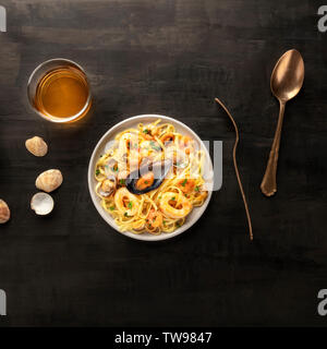 Piatti di pasta, overhead square shot. Tagliolini con cozze, gamberi, vongole e anelli di totano, con un bicchiere di vino bianco su sfondo nero Foto Stock