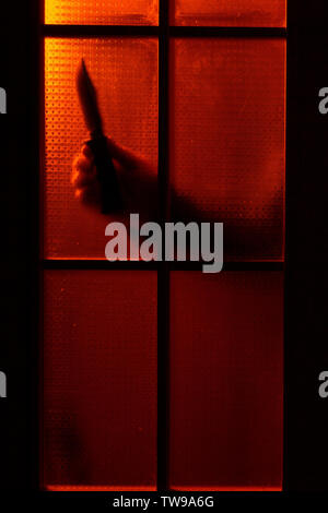 Mano d'uomo con un coltello dietro la porta con retroilluminazione rossa Foto Stock