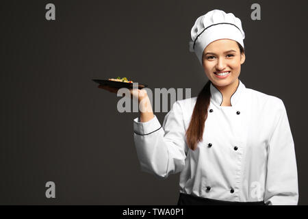 Bellissima femmina chef tenendo la piastra con piatto su sfondo scuro Foto Stock
