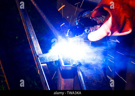 Saldatore lavoratore esegue la saldatura di salto. L'uomo saldatore in guanti di protezione esegue arc-procedimento di saldatura di strutture metalliche. Scintille volanti dalla saldatura Foto Stock