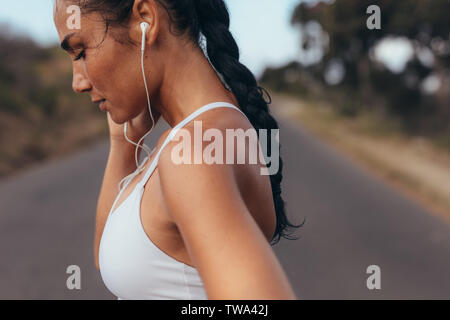 Vista laterale di un fitness donna in piedi su una strada vuota. Donna in usura fitness facendo Allenamento in piedi su una strada ascoltando la musica tramite gli auricolari. Foto Stock