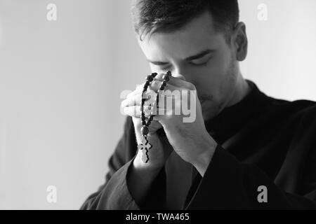 Giovane sacerdote con il Rosario pregando su sfondo chiaro, tonica in bianco e nero Foto Stock