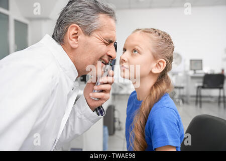 Vista dal lato del maschio oculista guardando attraverso lo strumento speciale alla pupilla degli occhi in clinica. Medico di sesso maschile di fare l'esame medico e controllando gli occhi della bambina in archivio ottico. Concetto di visione. Foto Stock