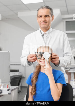 Vista frontale di felice ragazza con gli occhiali con lenti per il controllo della visione. Petty bambino chiudere occhio con la mano e guardando le lettere mentre ottico in piedi dietro in clinica. Concetto di Cura degli occhi. Foto Stock