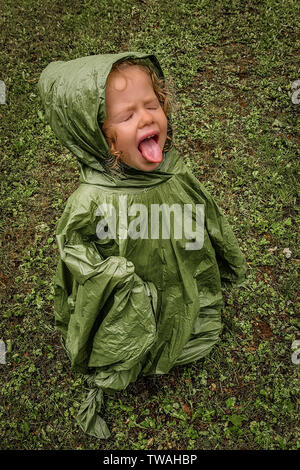 Poco toddler girl, giocando in pioggia, cercando di gusto, con gli occhi chiusi e la lingua di fuori, con verde cappotto di pioggia, su di un prato verde Foto Stock