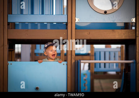 Little Boy, indossando il cappuccio, gridando a qualcuno, in piedi dietro alla parete, su un parco giochi di colore blu Foto Stock