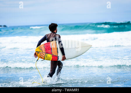 Surfer ragazzo con la tavola da surf in mano a correre verso grandi onde. Uomo in surf muta umida è in esecuzione in ondate di freddo oceano atlantico in Galizia, Spagna. Foto Stock