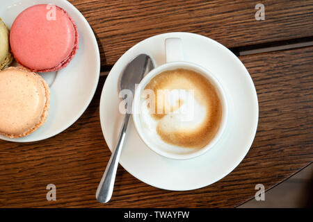 Caffè espresso caffè macchiato in ceramica bianca tazza con piattino e spon accanto a una piastra con macarons su marrone superficie di legno dal di sopra. Foto Stock