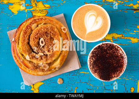 Due cappuccini e cannella rotolo di pasta isolati su peeling e incrinato blu e giallo legno dipinto dal di sopra. Foto Stock