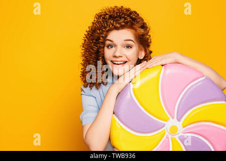 Ricci i capelli rossi positivo donna che indossa vestito blu con i dolci di grande lollipop su sfondo giallo Foto Stock